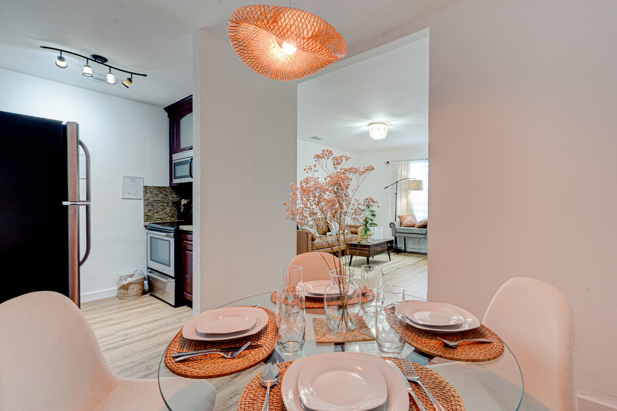 A dining area is depicted featuring a round glass table set for four, complete with dinner plates, glasses, and woven placemats. A light fixture with a woven design hangs above. Soft lighting and a glimpse of the living area can be seen in the background.