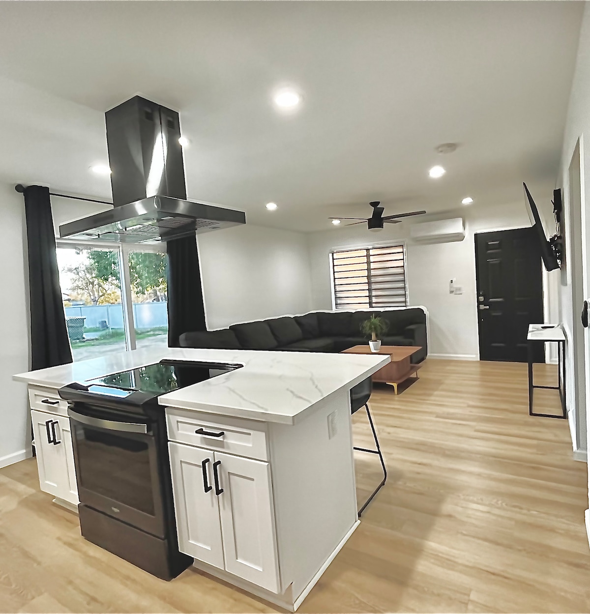 A modern kitchen and living area are integrated, featuring a central island with a stovetop and sleek cabinetry. A black sectional sofa is positioned near a large window, allowing natural light to fill the space. Decorative lighting and wooden flooring enhance the clean design.