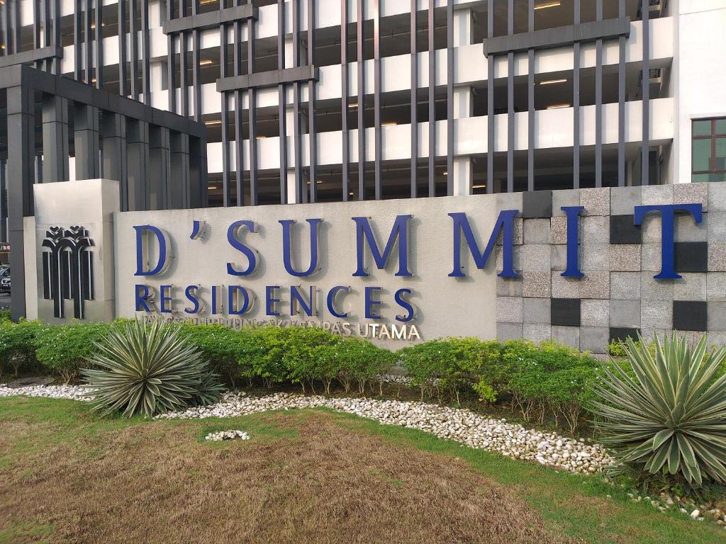 The entrance sign of D'Summit Residences is prominently displayed, featuring bold blue lettering against a stone background. Surrounding greenery includes well-maintained plants and shrubs, contributing to a welcoming ambiance. The structure of the building can be seen in the background, showcasing modern architectural elements.
