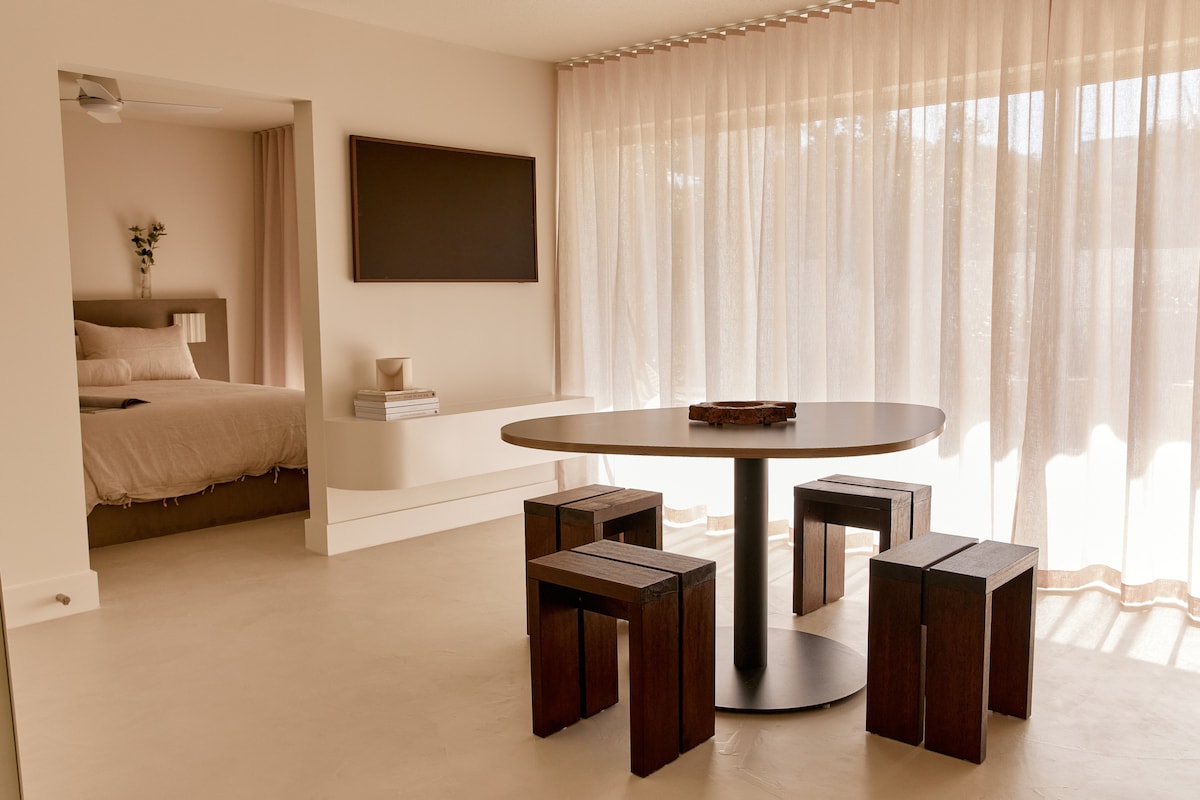 An open living area features a round dining table surrounded by four wooden stools. Light drapes softly through sheer curtains, illuminating the adjacent bedroom area, where a neatly made bed is visible. A flat-screen TV hangs on the wall opposite the table.