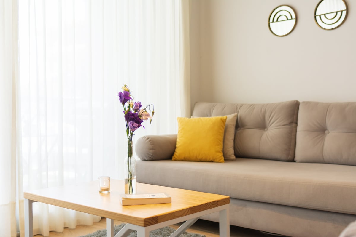 A light-filled living area features a neutral-toned sofa adorned with a bright yellow cushion. A wooden coffee table sits in front, holding a small book and a candle. Soft, sheer curtains gently filter sunlight, contributing to the serene ambiance.
