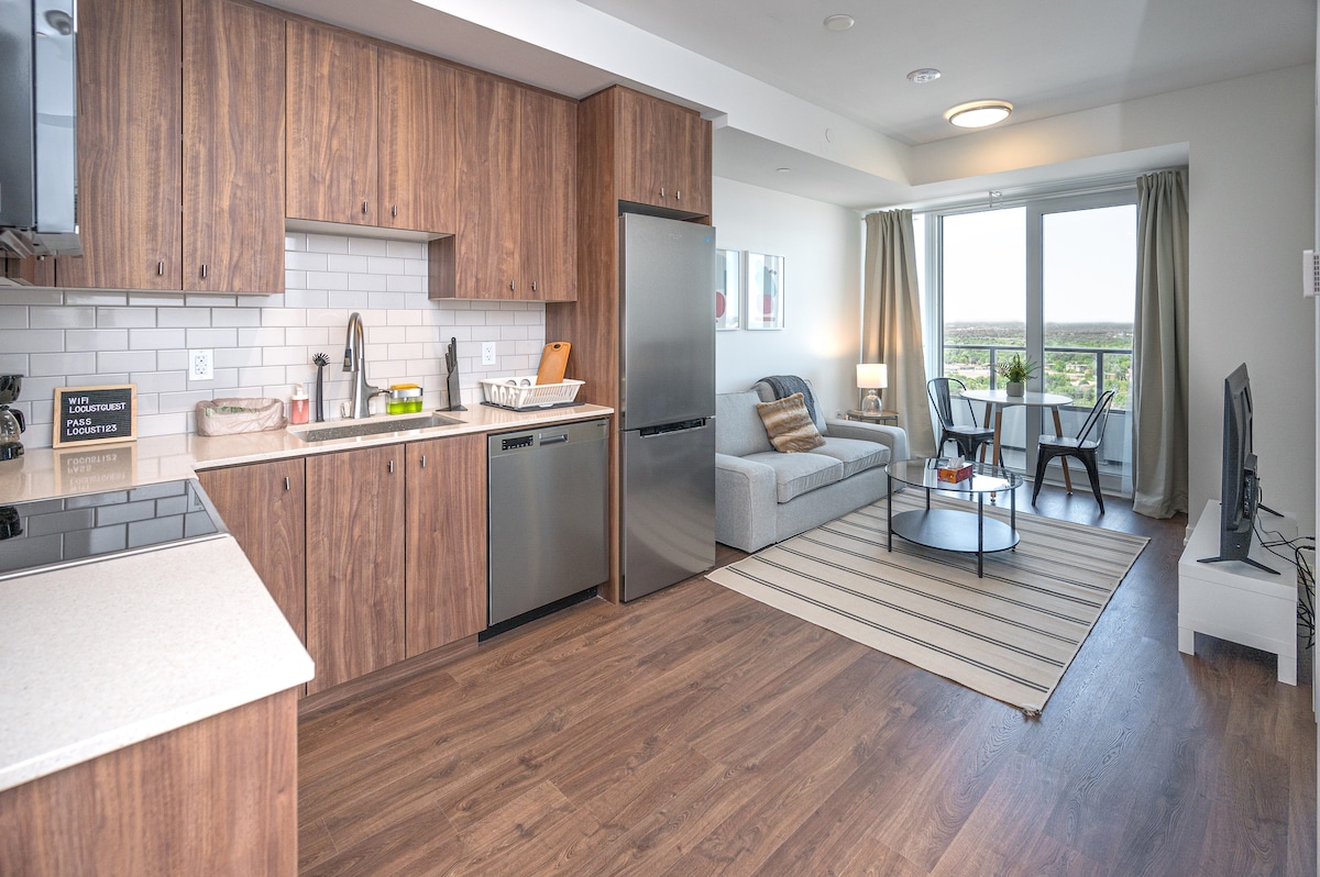 A modern kitchen features wooden cabinetry and stainless-steel appliances, including a refrigerator and dishwasher. A cozy living area is visible, equipped with a comfortable sofa and a coffee table on a striped area rug. Natural light fills the space through large glass doors that lead to a balcony.