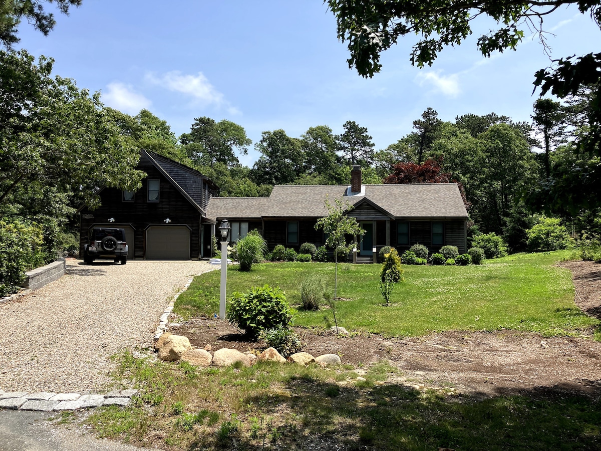 A spacious home exterior is surrounded by well-maintained landscaping and a gravel driveway. The two-story structure features a combination of dark wood siding and light-colored roofing. Large windows provide ample natural light. Lush greenery and decorative stones enhance the welcoming facade.