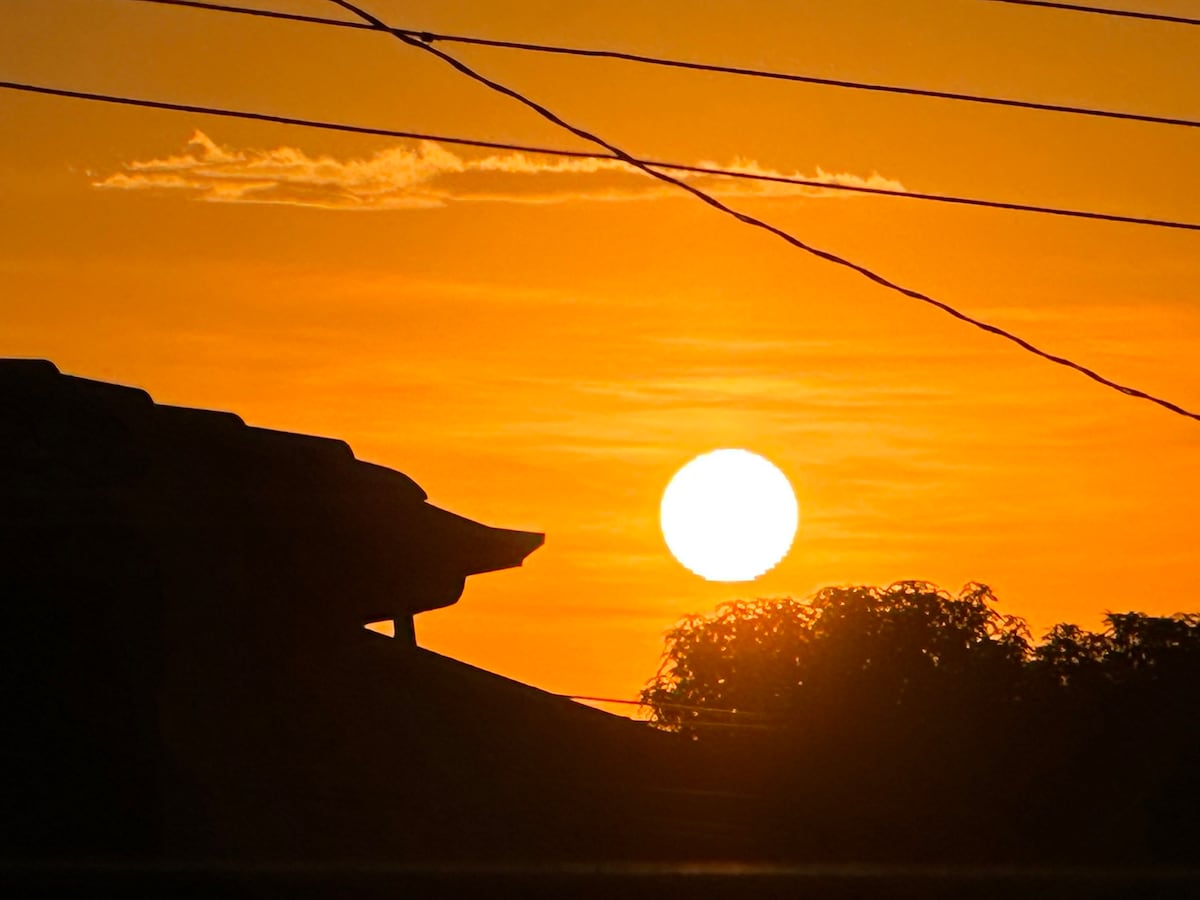 A warm sunset is captured against a vibrant orange sky, with the sun appearing large and round. Silhouetted rooftops and treetops create a peaceful horizon, while power lines subtly intersect the image.