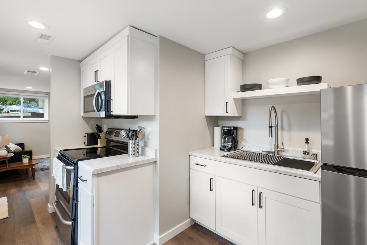 A modern kitchen showcases sleek white cabinetry and marble countertops. Stainless steel appliances, including an oven and refrigerator, are visible. A sink with a stylish faucet is positioned next to a coffee machine. Ample natural light filters through a nearby window.