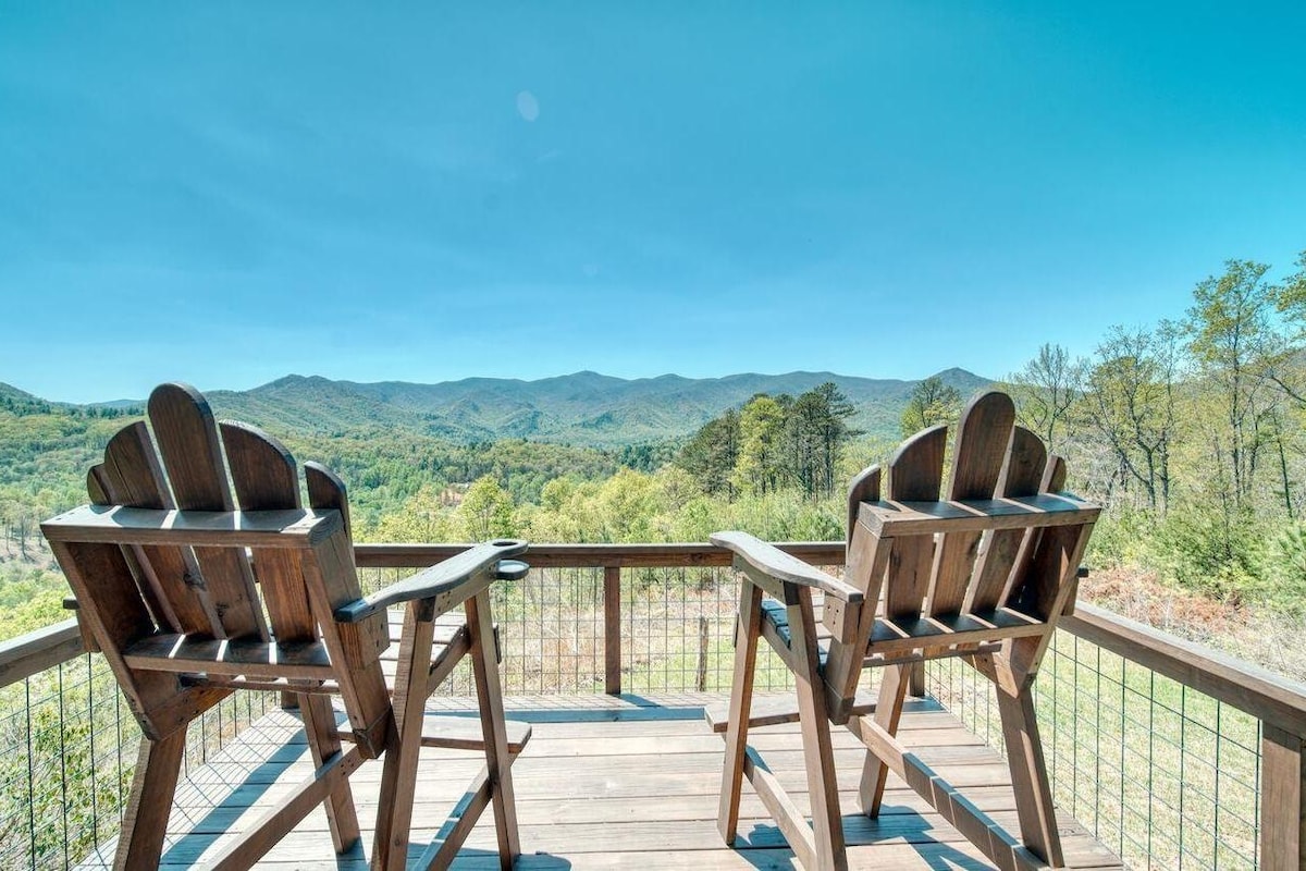 A spacious outdoor deck features two wooden adirondack chairs, positioned to provide unobstructed views of rolling mountains and lush greenery. The clear sky contributes to the bright atmosphere, creating a serene environment for relaxation.