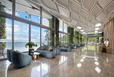 The lobby features expansive glass windows offering a view of the sea. Plush seating is arranged to encourage relaxation, complemented by indoor plants. Natural light fills the space, enhancing the modern design with geometric ceilings and elegant marble flooring.