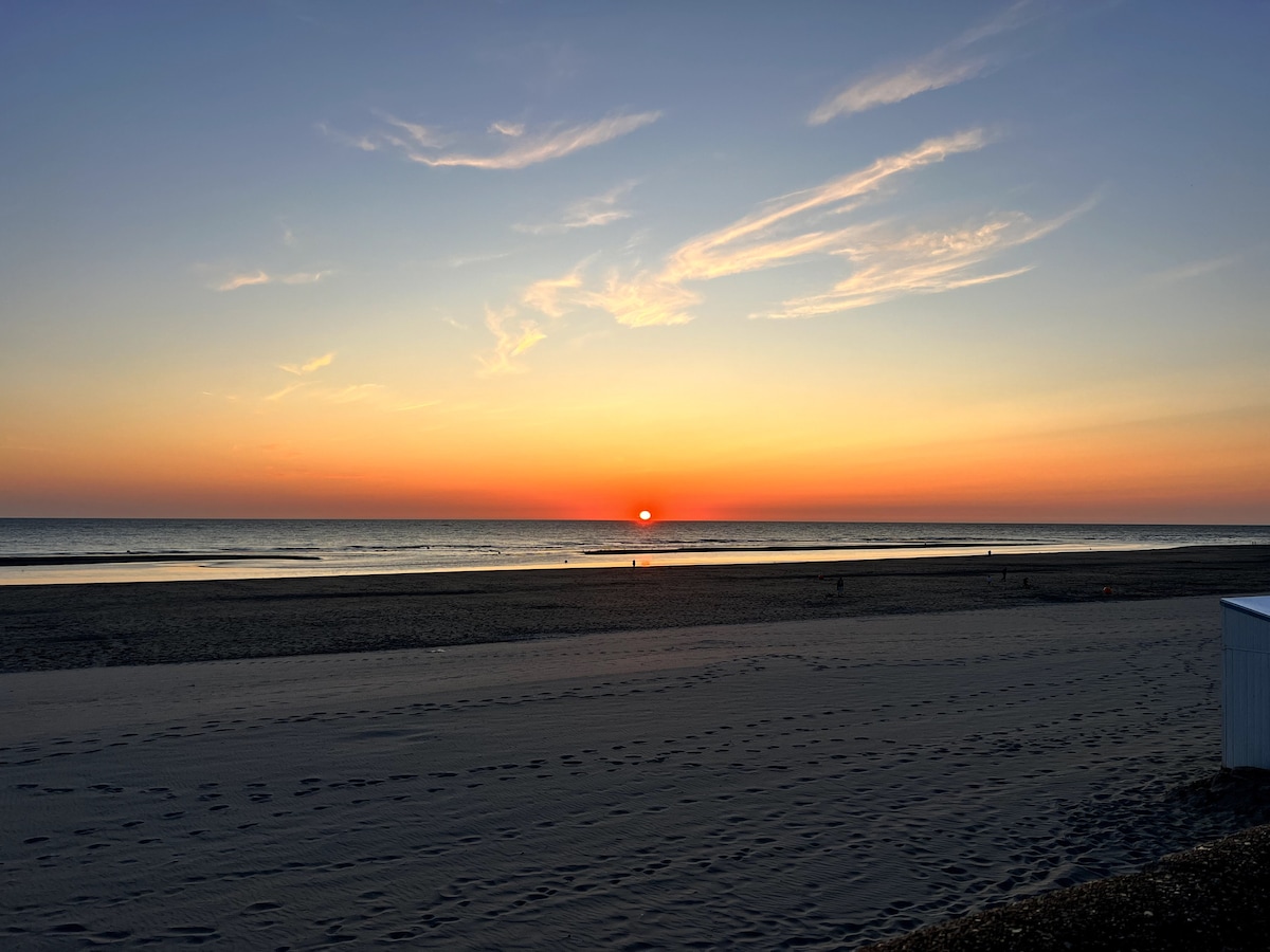 A serene beach scene captures the setting sun, casting warm hues of orange and pink across the sky. The calm ocean waves reflect the colorful sky as footprints in the sandy foreground lead towards the water's edge.