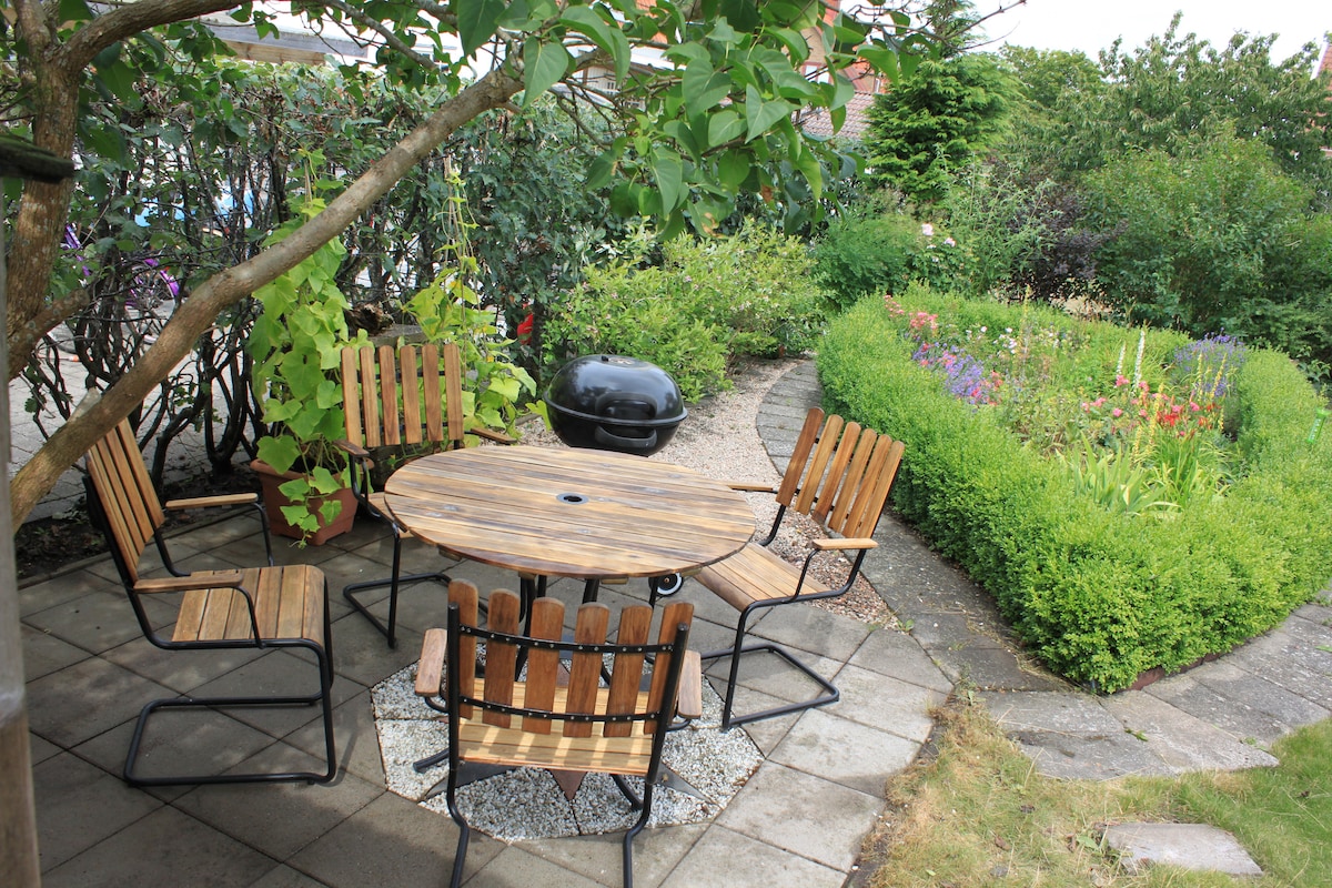 An outdoor seating area features a round wooden table surrounded by six chairs, positioned on a stone-paved surface. Lush greenery and colorful flower beds create a serene garden backdrop, while a small barbecue grill is placed nearby.