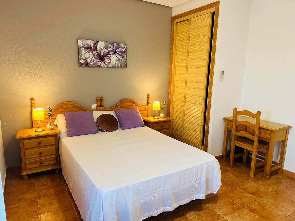 A bright bedroom features a spacious bed with white linens and purple accent pillows. On either side of the bed, wooden nightstands hold softly glowing lamps. A wooden desk is visible, with a floral wall art piece above the bed. Light-colored walls and floor tiles enhance the room's openness.