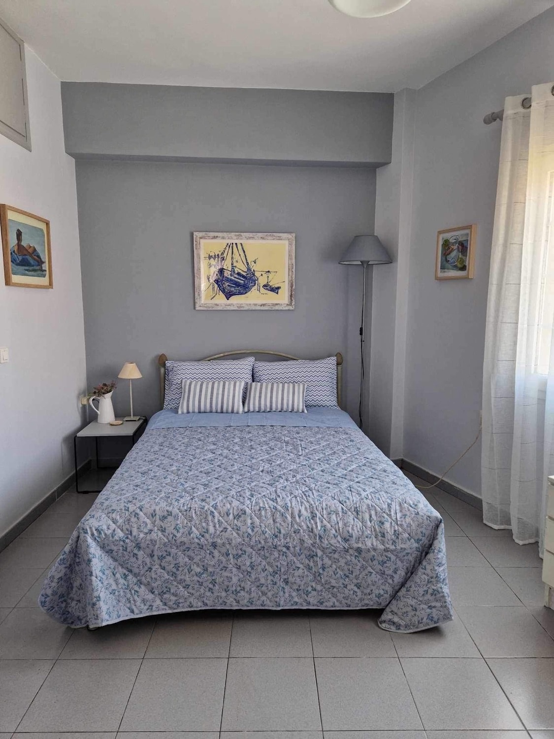 A neatly arranged sleeping area features a double bed covered with a patterned blue quilt. Two striped pillows are placed against the headboard. A bedside lamp and a small nightstand are positioned beside the bed. Soft natural light filters through the sheer curtains.