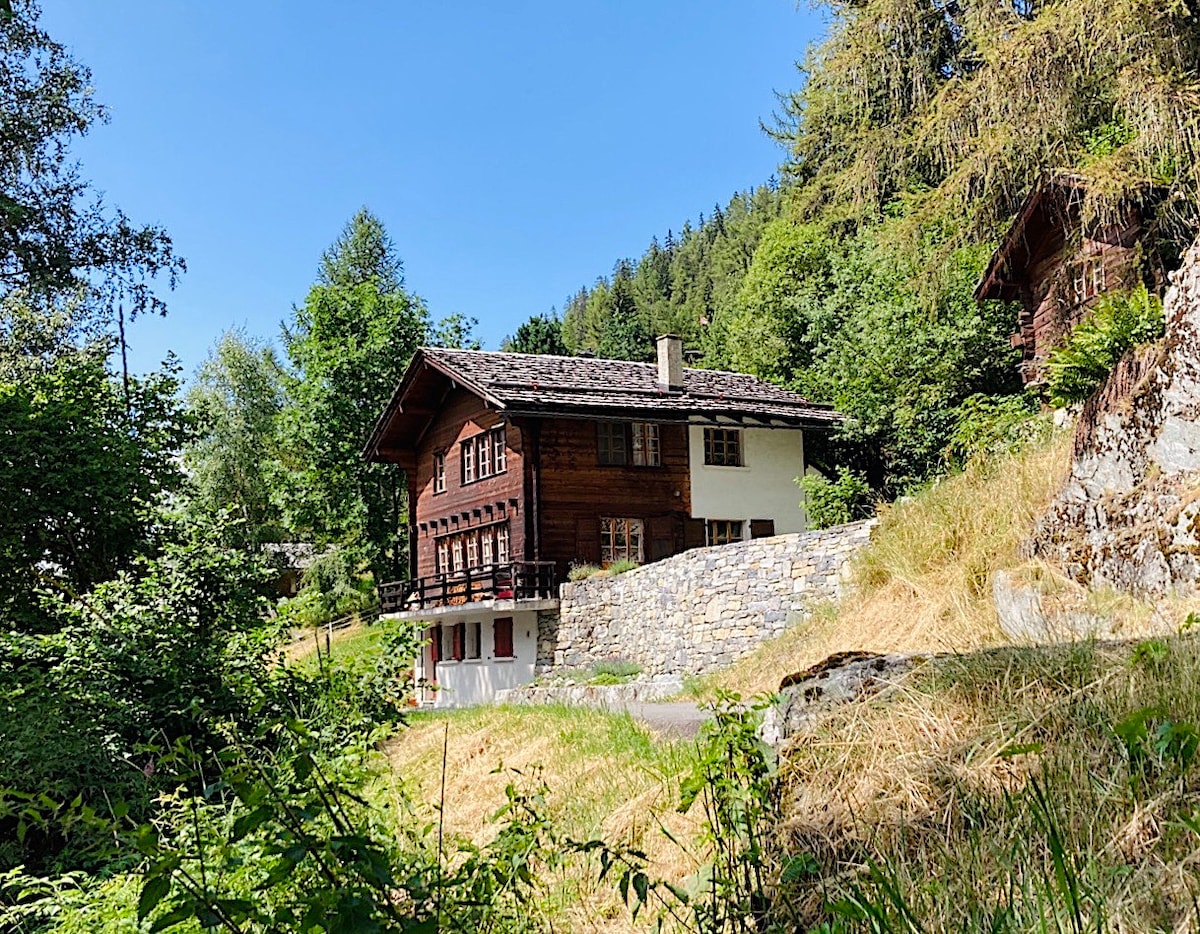 A charming chalet is nestled among greenery, surrounded by lush trees and grassy areas. The building features traditional wooden architecture with a sloped roof and stone accents. Sunny skies illuminate the exterior, enhancing the inviting atmosphere of this tranquil retreat.