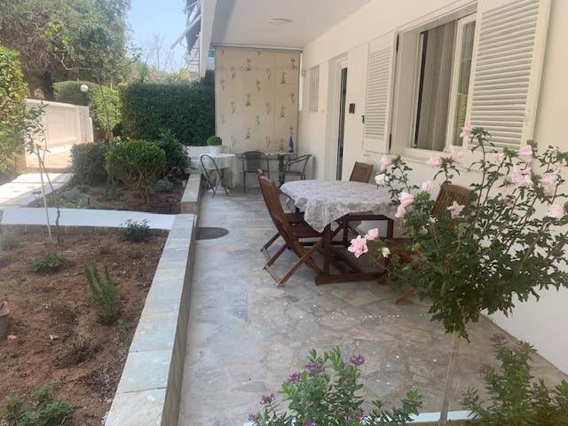 An outdoor terrace features a wooden dining table surrounded by folding chairs, with a light-colored tablecloth on top. Flanking the terrace are well-maintained flower beds and shrubs, enhancing the natural surroundings. The entrance to the studio is visible in the background.