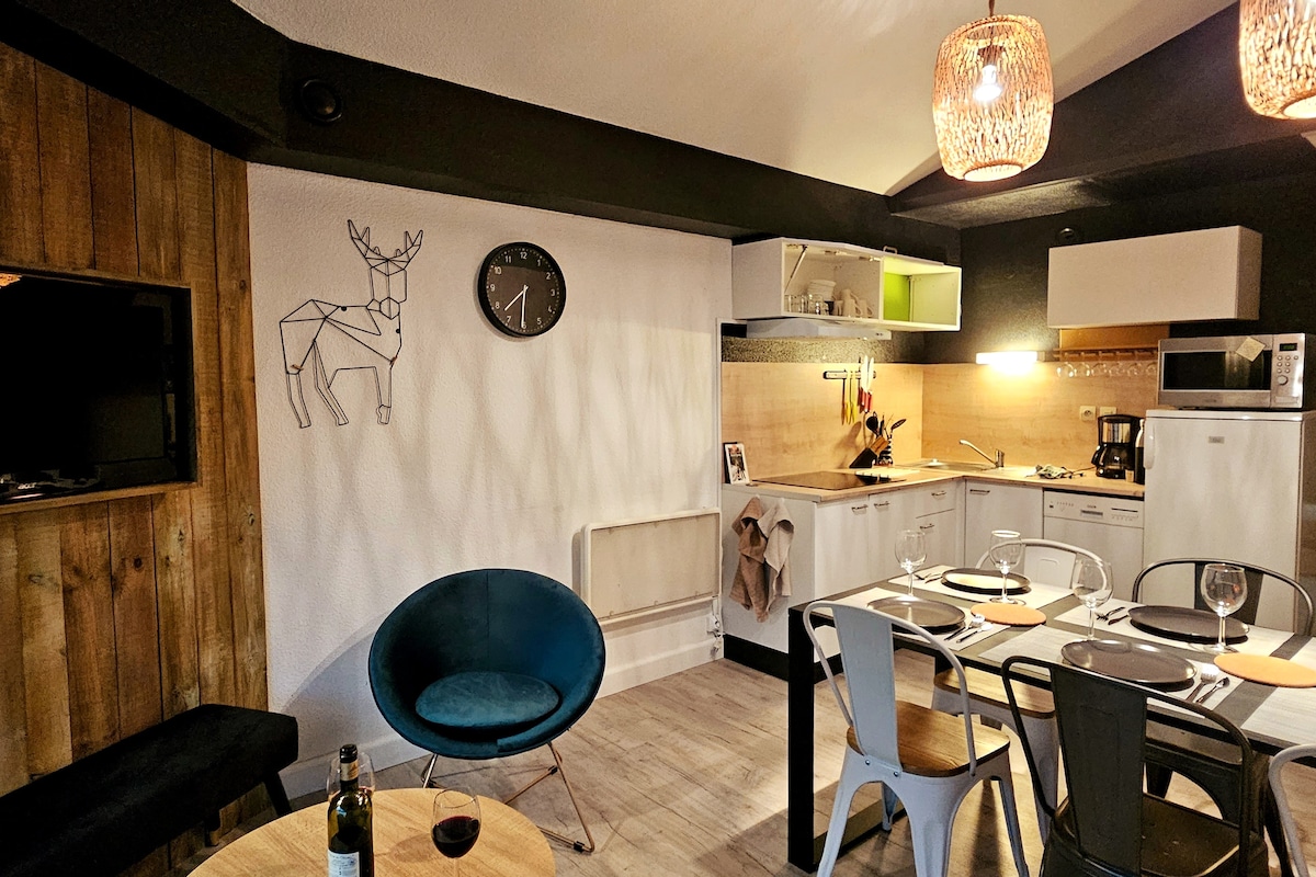 A modern kitchen area features sleek cabinetry and a dining table set for four. A decorative clock and wall art add charm to the space. Soft lighting is provided by woven pendant lamps, and a cozy seating arrangement includes a blue chair and a small table.