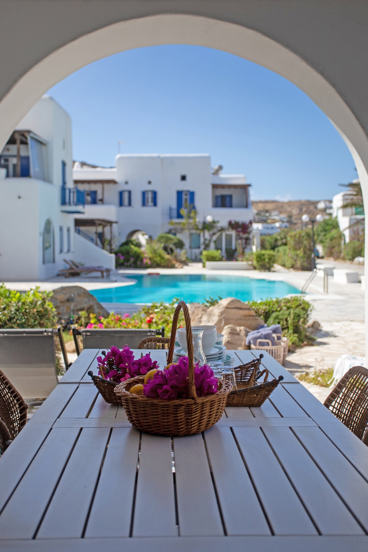 An outdoor dining area is framed by an archway, featuring a white table set with a basket of bread and vibrant pink flowers. A shared swimming pool is visible in the background, surrounded by lush greenery, with white buildings in the distance under a clear blue sky.