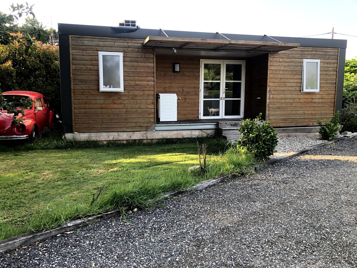 A modern wooden structure features a covered front porch with glass doors. Two windows are aligned symmetrically on either side of the entrance. Lush greenery surrounds the exterior, complemented by a classic red car parked nearby on a gravel path.