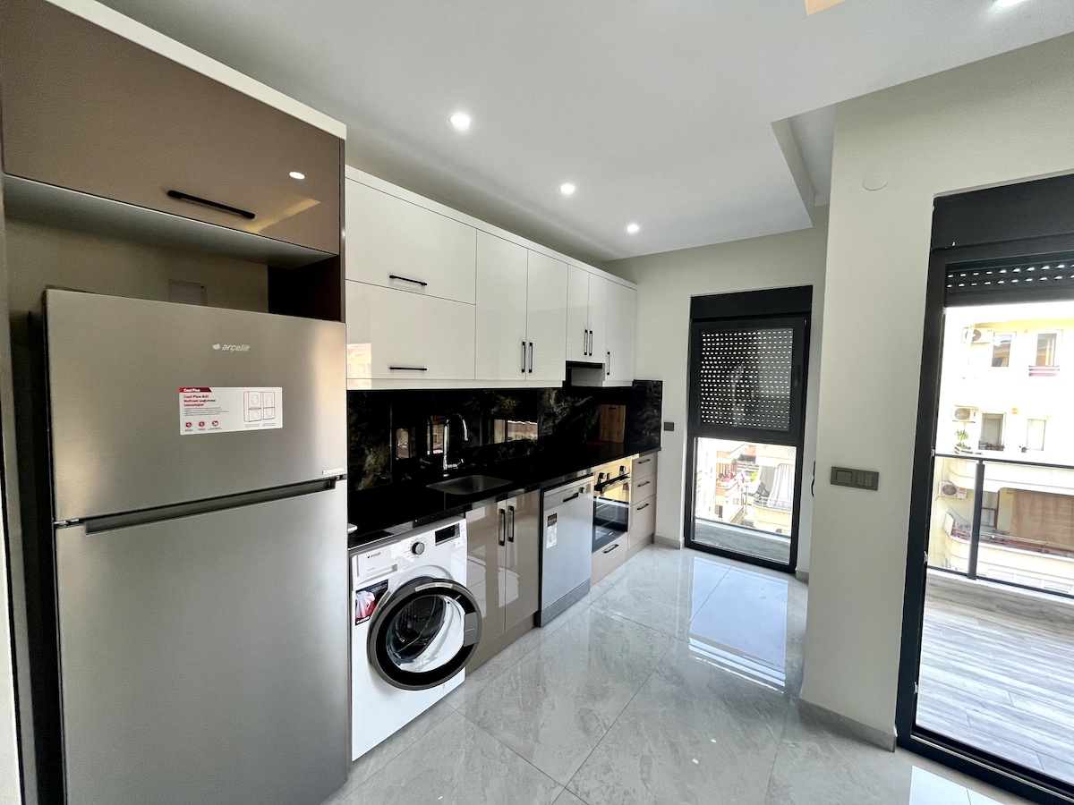 A modern kitchen is presented with sleek cabinetry and a stylish refrigerator. Countertop appliances are positioned alongside a washing machine and oven, while black marble surfaces enhance the overall design. A window provides natural light and displays views of the surrounding area.