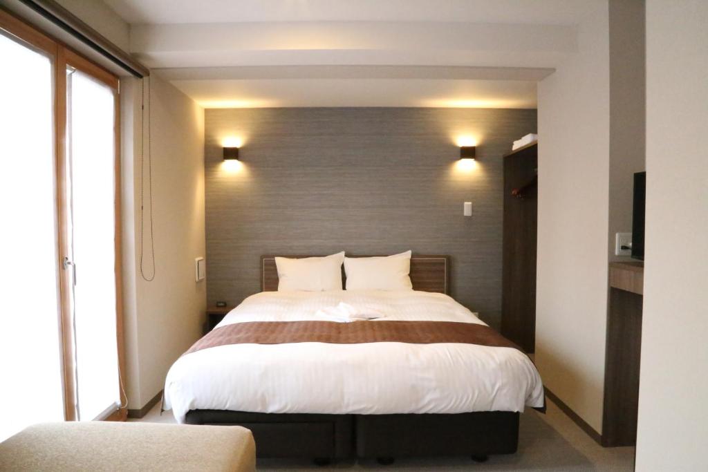 A spacious sleeping area is furnished with a large bed, featuring white bedding and brown accents. Soft lighting from wall sconces creates a calming environment. The room includes ample natural light from glass doors leading to a small balcony or outdoor space.