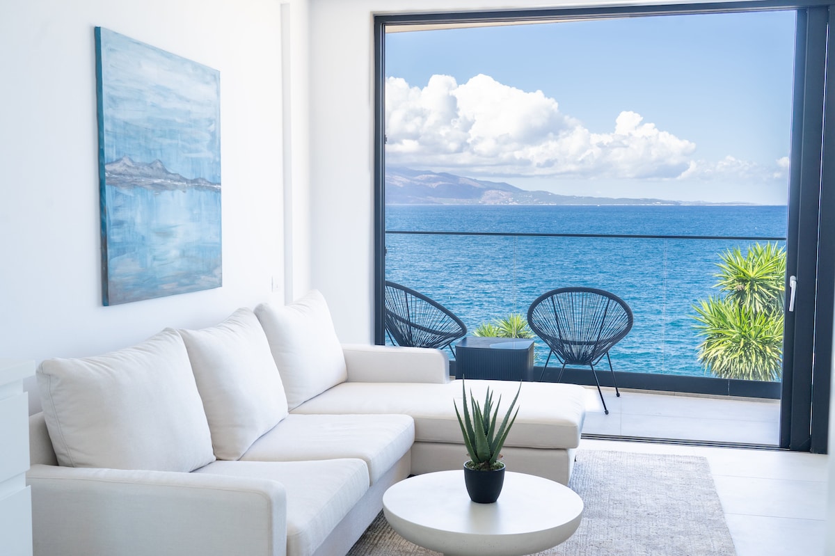 A bright living area features a light-colored sectional sofa paired with a small round table. Large glass doors provide a view of the sea and distant mountains, while two outdoor chairs and palm plants are visible on the balcony.