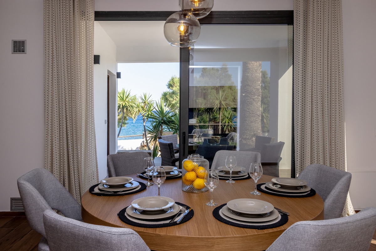 A round dining table is set for a meal, featuring elegant dishware and glasses. Sunlight filters through large glass doors, providing views of the lush palm trees and the Aegean Sea beyond. Soft gray chairs surround the table, complementing the natural wood finish.