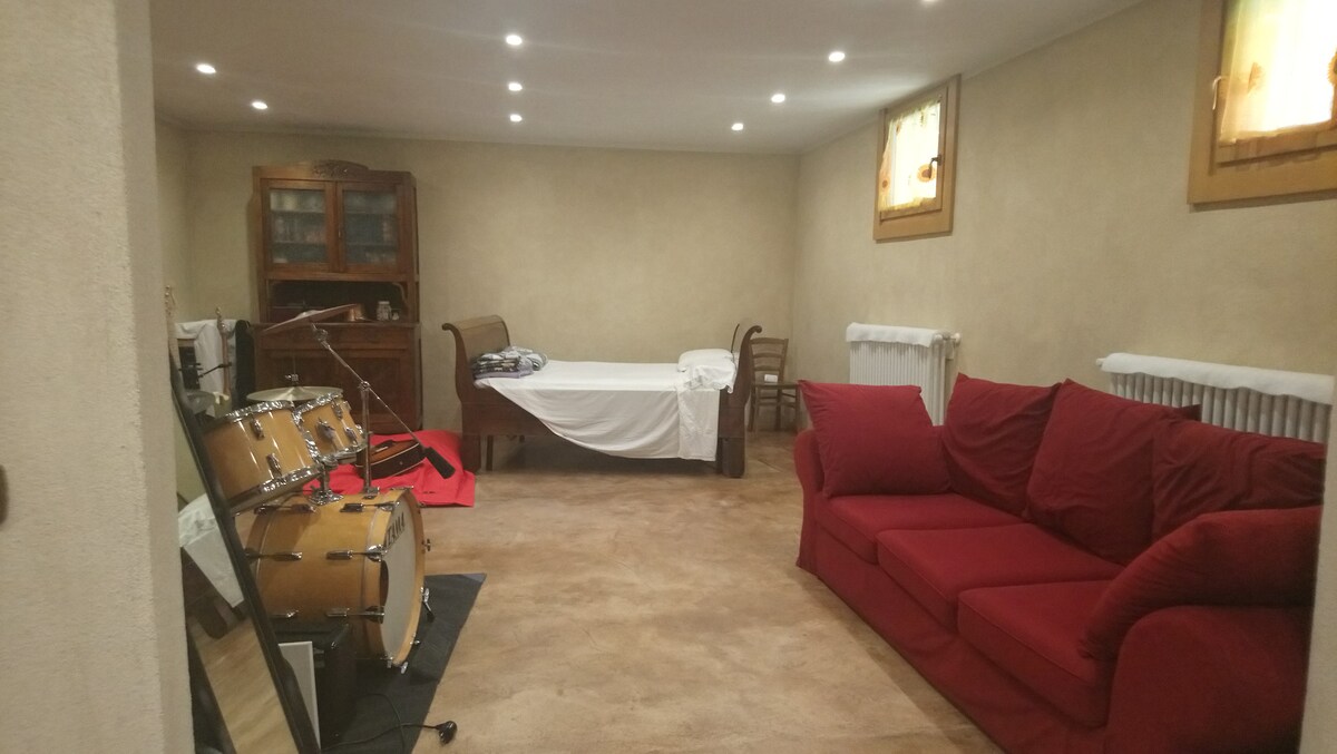 A spacious room features soft beige walls and a textured carpet. A red sofa is positioned next to a small wooden table, while a simple bed is covered with white linens. Drums are set up on a mat, and three windows allow natural light to enhance the area.