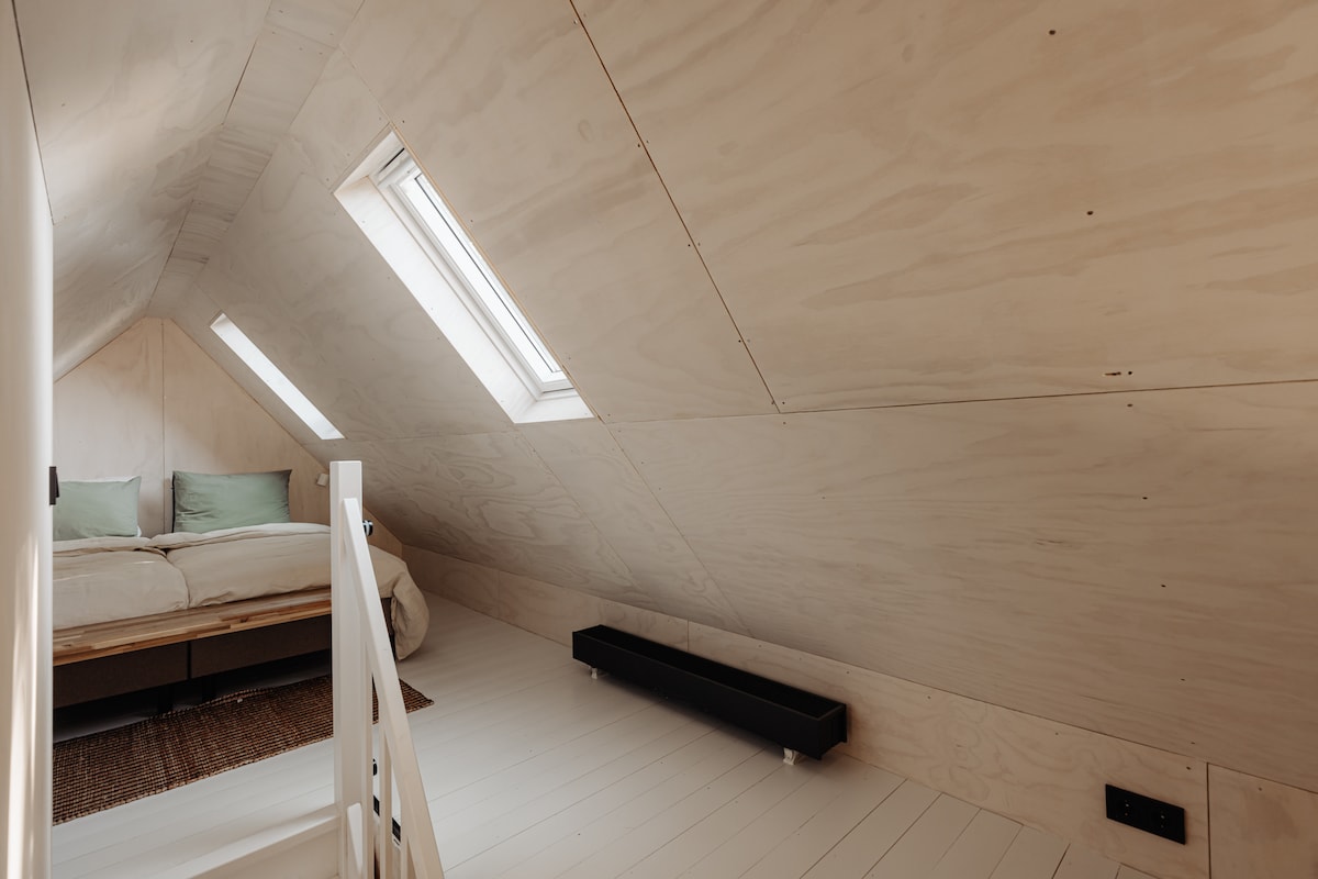 A cozy sleeping area is situated beneath sloped wooden walls, featuring a spacious bed adorned with soft green cushions. Natural light enters through skylights, illuminating the light-colored hardwood floor. A simple black bench is placed along one wall, contributing to the minimalist design.