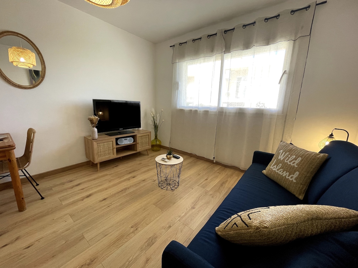 A comfortable living space features a dark blue sofa adorned with a decorative pillow. A round coffee table sits in front of the sofa, complemented by a wooden side table. Natural light filters through sheer curtains covering a large window, with a television positioned on a cabinet across the room.