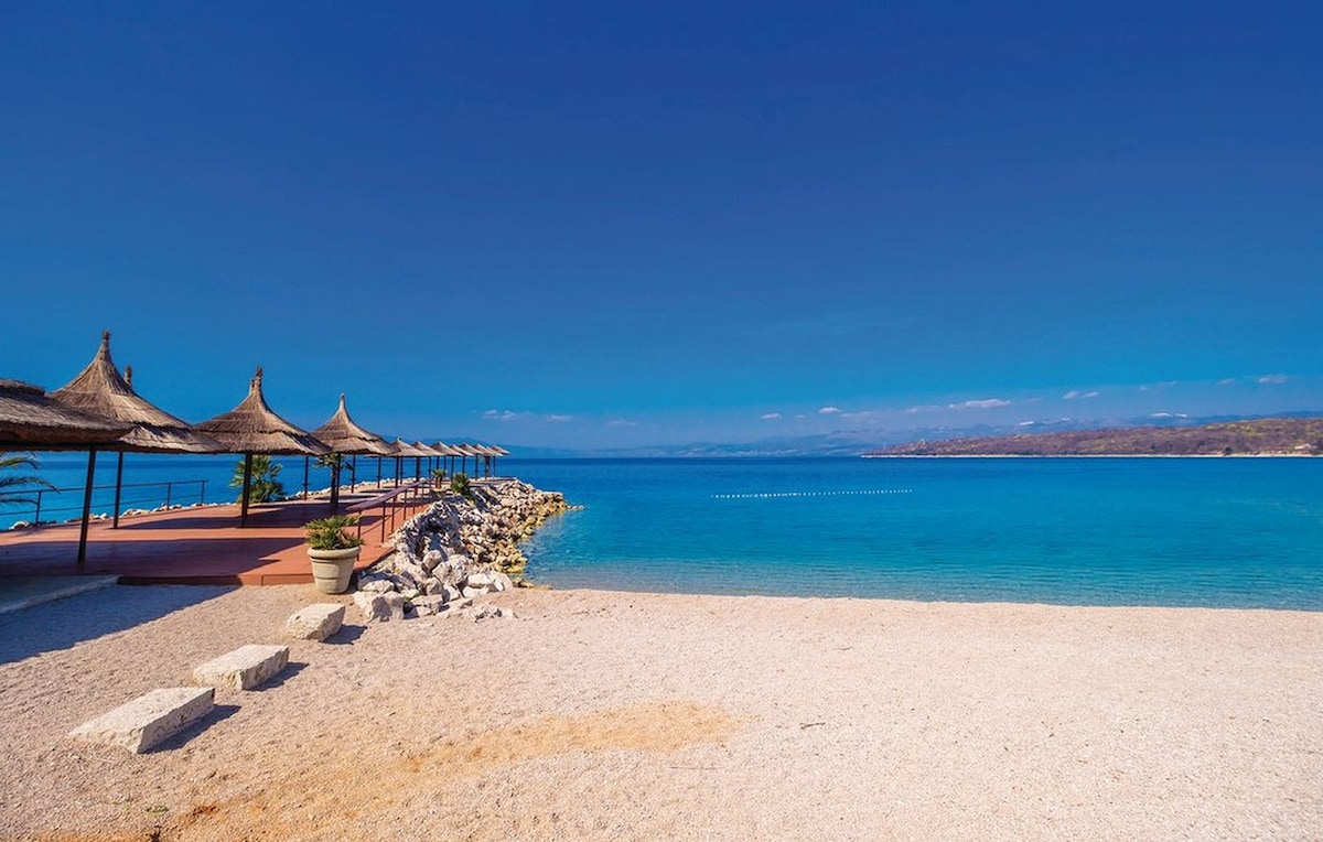 A serene beachfront is presented, featuring soft pebbles and clear turquoise waters. Thatched roof sunshades line the pathway, providing shaded areas for relaxation. The calm sea stretches into the horizon, with distant land visible against a clear blue sky.