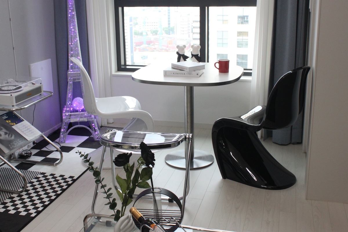 A modern seating area shows a round table flanked by sleek black and white chairs. A small Eiffel Tower decoration stands beside the window, which offers views of the cityscape. A decorative tray holds two wine glasses and a bottle, adding a touch of elegance.