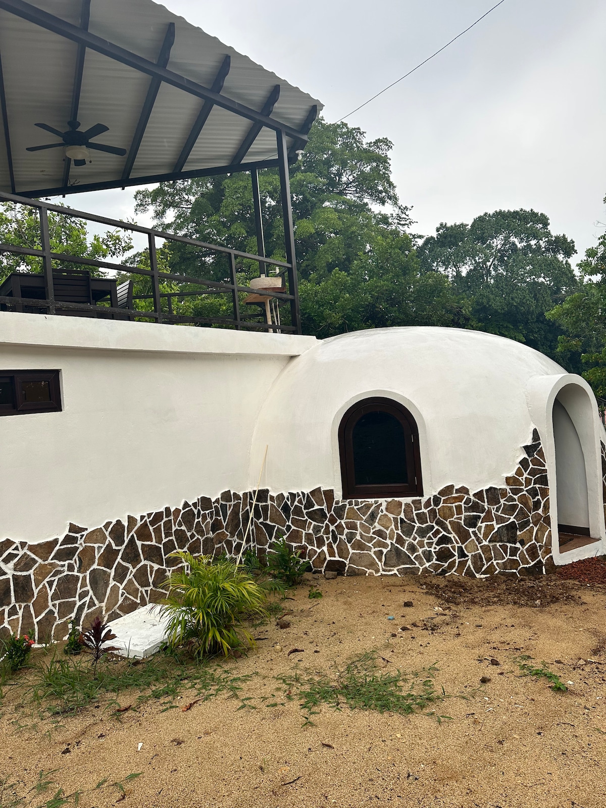 A unique architectural structure featuring a rounded white dome and a stone accent wall. An outdoor deck with seating is visible above the dome, surrounded by greenery. The setting offers a natural, earthy feel, inviting connections with the surrounding environment.