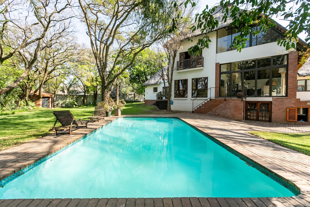 A clear turquoise pool is set among a spacious garden featuring large trees. Lounge chairs are positioned on the deck near the water's edge. The two-story residence with large windows and a welcoming entrance overlooks the pool area, enhancing the outdoor retreat.