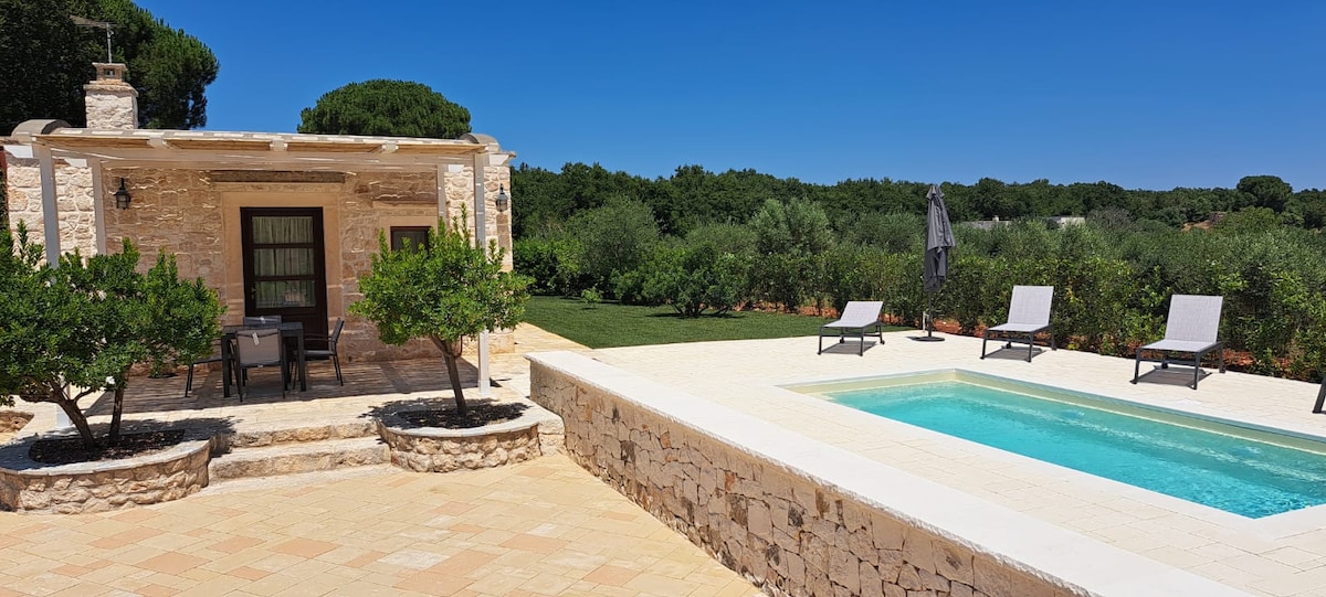 A villa features a private pool surrounded by sun loungers and an umbrella. A patio with seating is adjacent to the pool, framed by landscaped greenery in the background. The exterior is constructed with light-colored stone and offers a serene outdoor space.