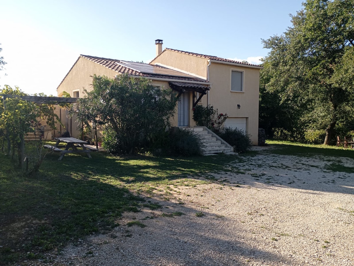 A rural house is surrounded by greenery, featuring a covered porch area. Stone steps lead up to the main entrance. The gravel driveway is framed by grass, and trees provide shade nearby, creating a serene outdoor setting.