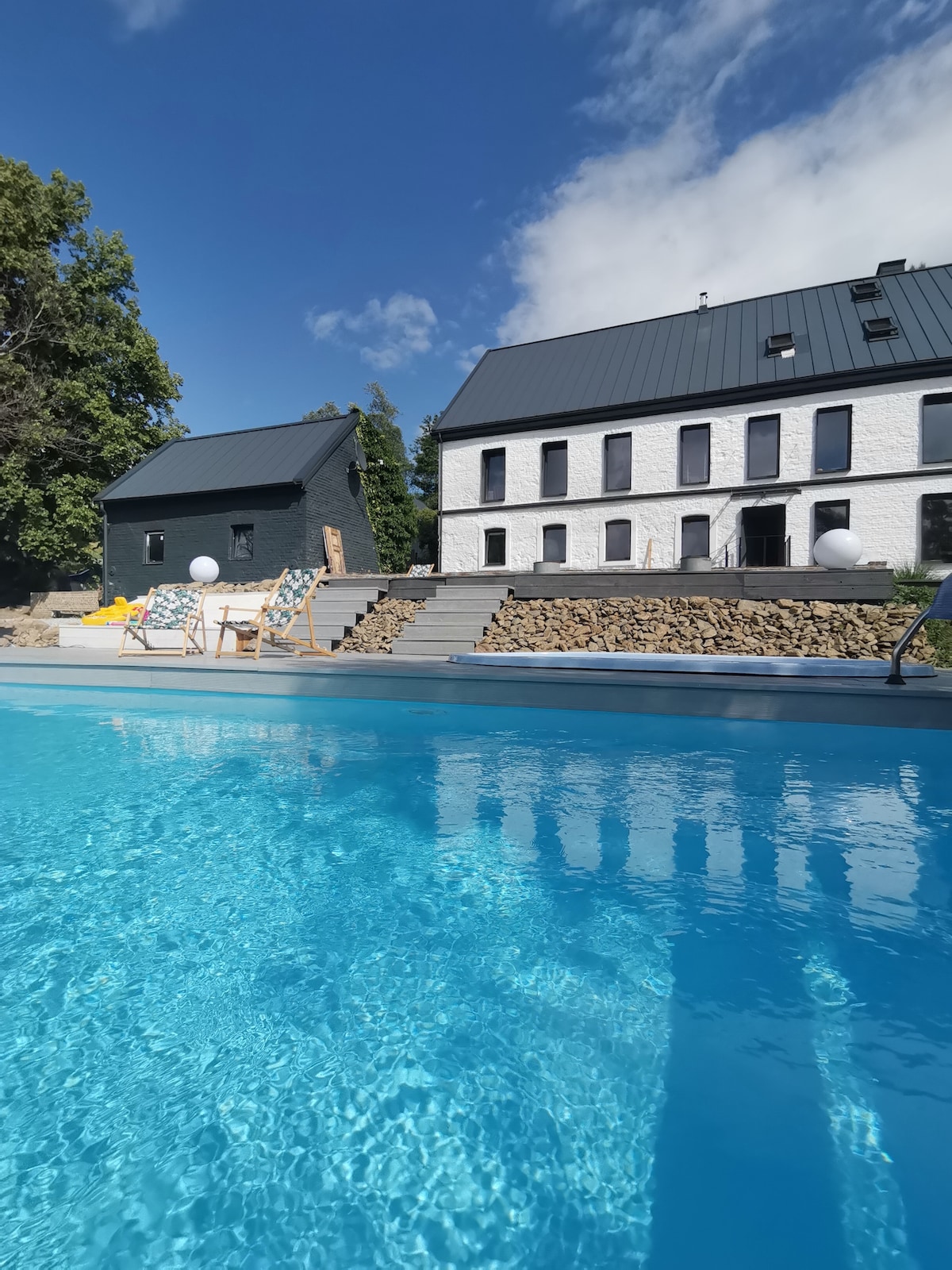 A clear view of a large, heated swimming pool reflects the blue sky. The pool area is surrounded by landscaped stone steps leading to stylishly designed buildings with dark roofs and white walls. Sun loungers provide a spot for relaxation beside the water.