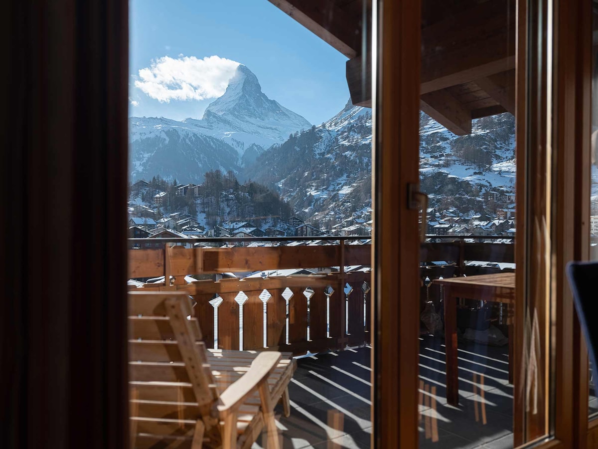 A scenic view from a balcony showcases a snow-covered mountain, with expansive glass doors allowing natural light to fill the space. Outdoor seating is positioned on the deck, complemented by wooden railings that frame the picturesque landscape.