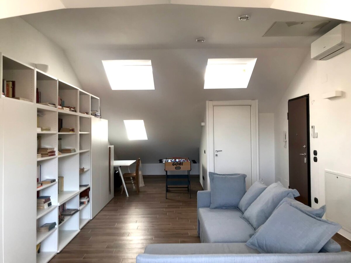An airy attic space features two skylights that allow natural light to fill the room. A cozy seating arrangement includes a neutral-toned sofa. A well-stocked bookshelf lines one wall, while a foosball table is positioned nearby. A small workspace with a table awaits in the corner.