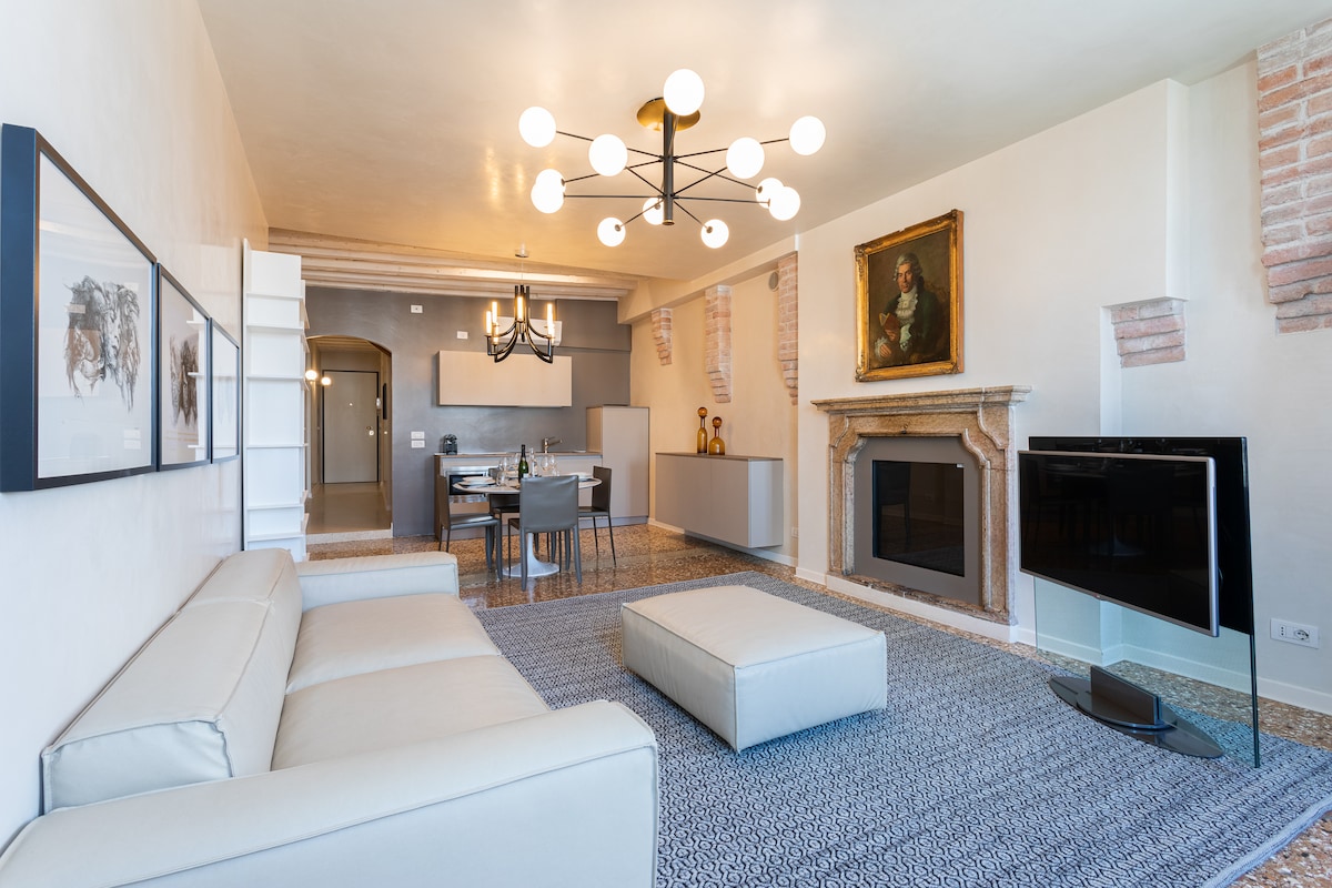 A modern living area features a neutral-colored sofa and large flat-screen television. A decorative area rug underfoot complements the light-colored walls and exposed brick accents. A dining table, set for guests, is visible in the background, alongside contemporary lighting fixtures that enhance the ambiance.