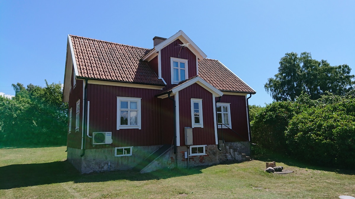 A charming red wooden house is featured, set against a clear blue sky. The structure showcases a steep roof, white window frames, and a small staircase leading to the entrance. Lush greenery surrounds the house, providing a natural backdrop.