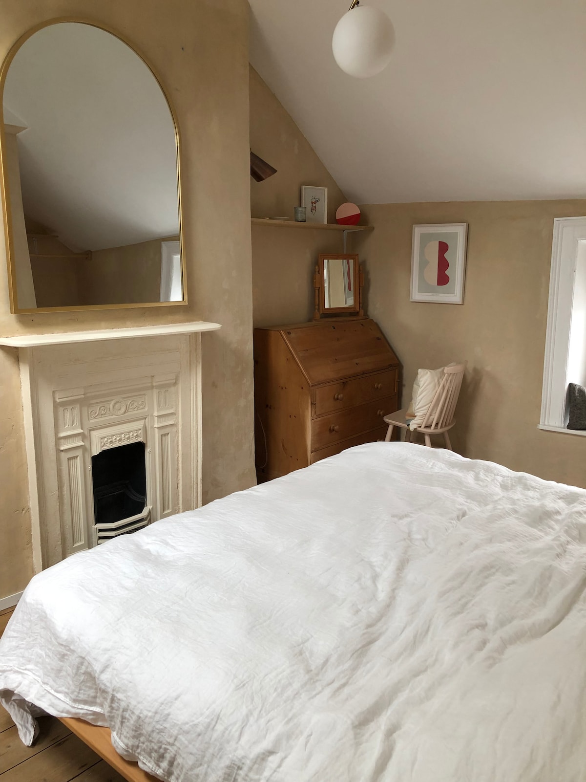 A light and airy bedroom features a spacious bed covered with a soft white duvet. An ornate mirror is positioned above a decorative fireplace, and light wood furnishings include a dresser and a chair. Subtle wall art complements the warm-toned walls.