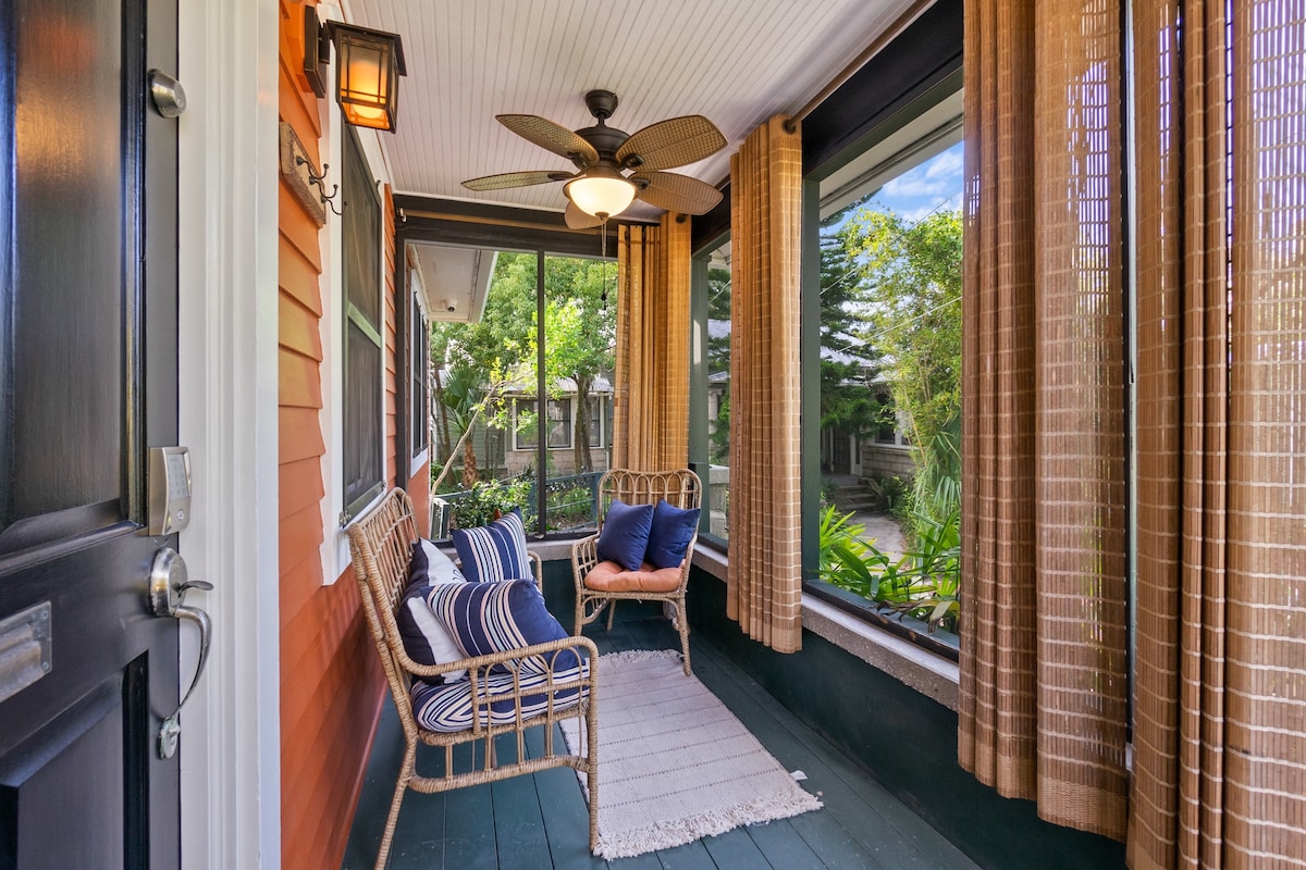 A serene screened-in porch features a comfortable loveseat and a pillowed chair, surrounded by large windows with natural light. Decorative curtains and a ceiling fan enhance the tranquil atmosphere, while lush greenery is visible in the outdoor area beyond.