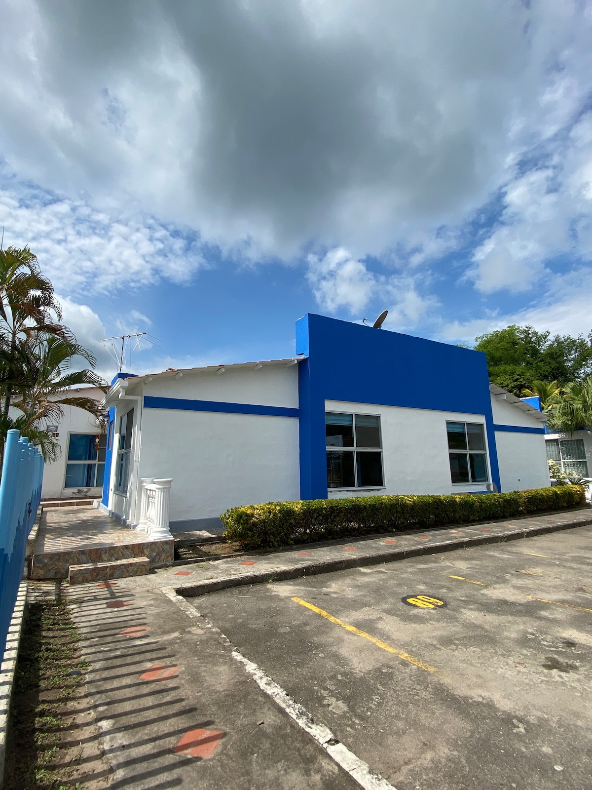The exterior of Villa Elisa is showcased, featuring a white structure with a bright blue accent wall. Lush plants border the property, with a paved pathway leading to the entrance. A clear blue sky with scattered clouds adds to the serene atmosphere.