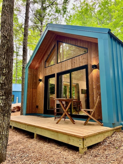 A modern cabin exterior is framed by tall trees, featuring a peaked roof and large glass doors. A wooden deck holds two folding chairs, providing a quiet spot to enjoy the natural surroundings.