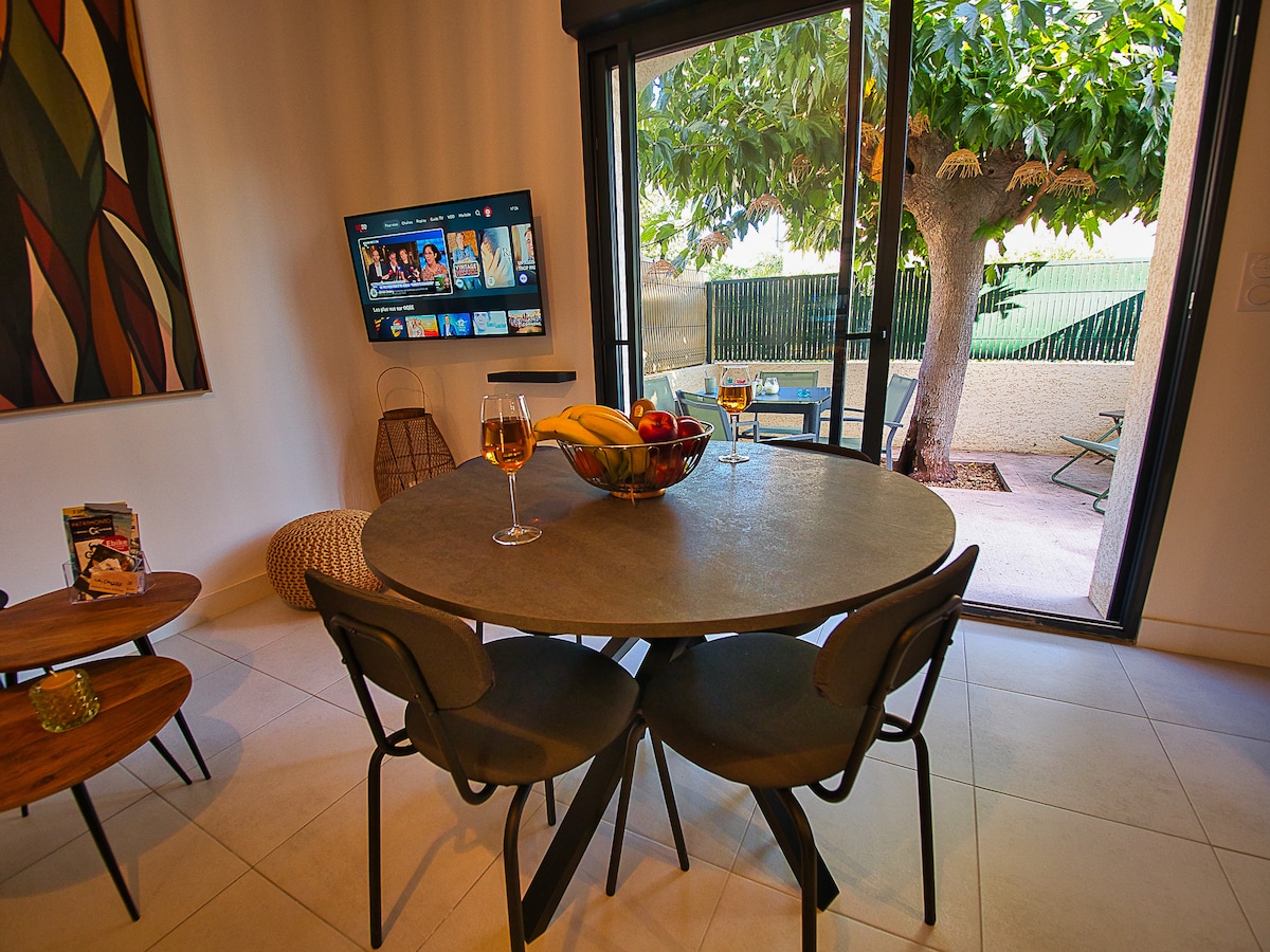 A modern dining area features a round stone table surrounded by four chairs. A large television is mounted on the wall, and large glass doors open to a patio area with outdoor seating. A fruit bowl is placed at the center of the table.