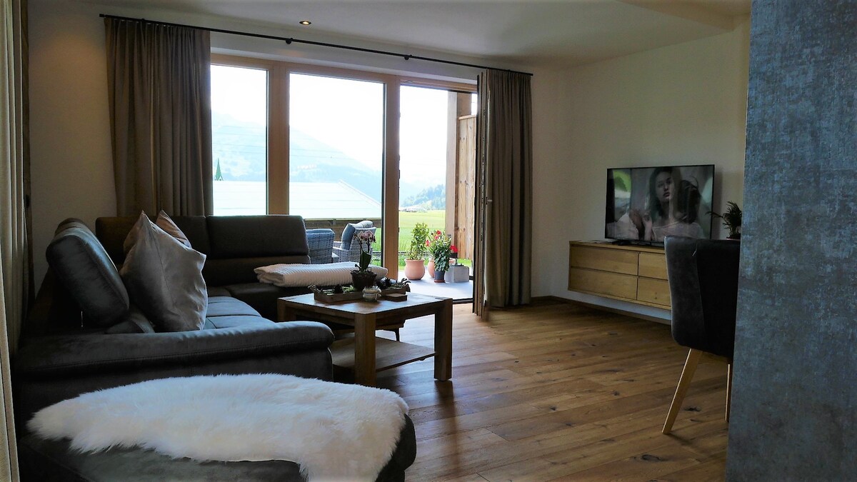 A spacious living area features a comfortable dark sectional sofa with soft cushions and a coffee table. Large windows open to a view of the surrounding landscape, allowing natural light to illuminate the space. A flat-screen TV is positioned on a wooden cabinet.