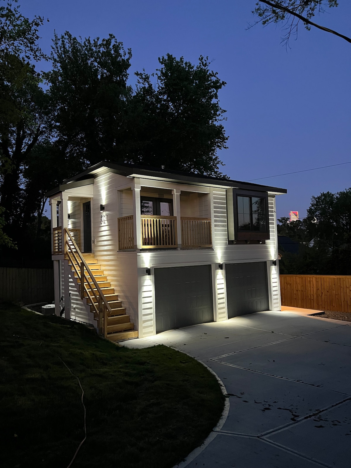 A charming two-story cottage is illuminated at dusk, highlighting the exterior's white siding and balcony. Soft lighting accents the entrance steps and garage doors, while surrounding greenery provides a serene backdrop. The private setting is enhanced by a a wooden fence enclosing the yard.