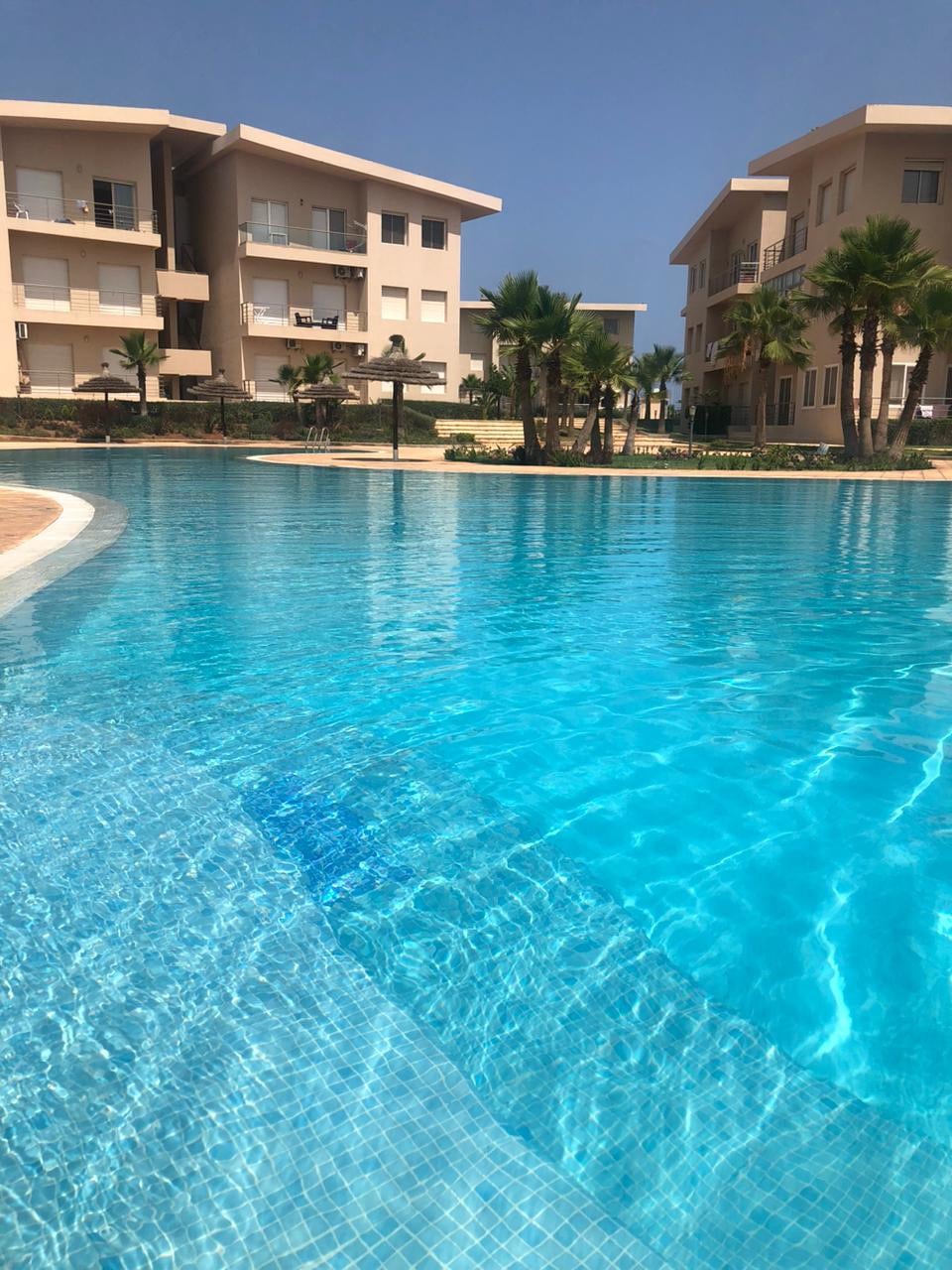 A clear and inviting swimming pool is shown with shimmering turquoise water reflecting the sunlight. Surrounding the pool, modern residential buildings and palm trees create a tranquil setting, emphasizing a resort-like atmosphere. The pool’s edge features a smooth transition from the water to the surrounding architecture.