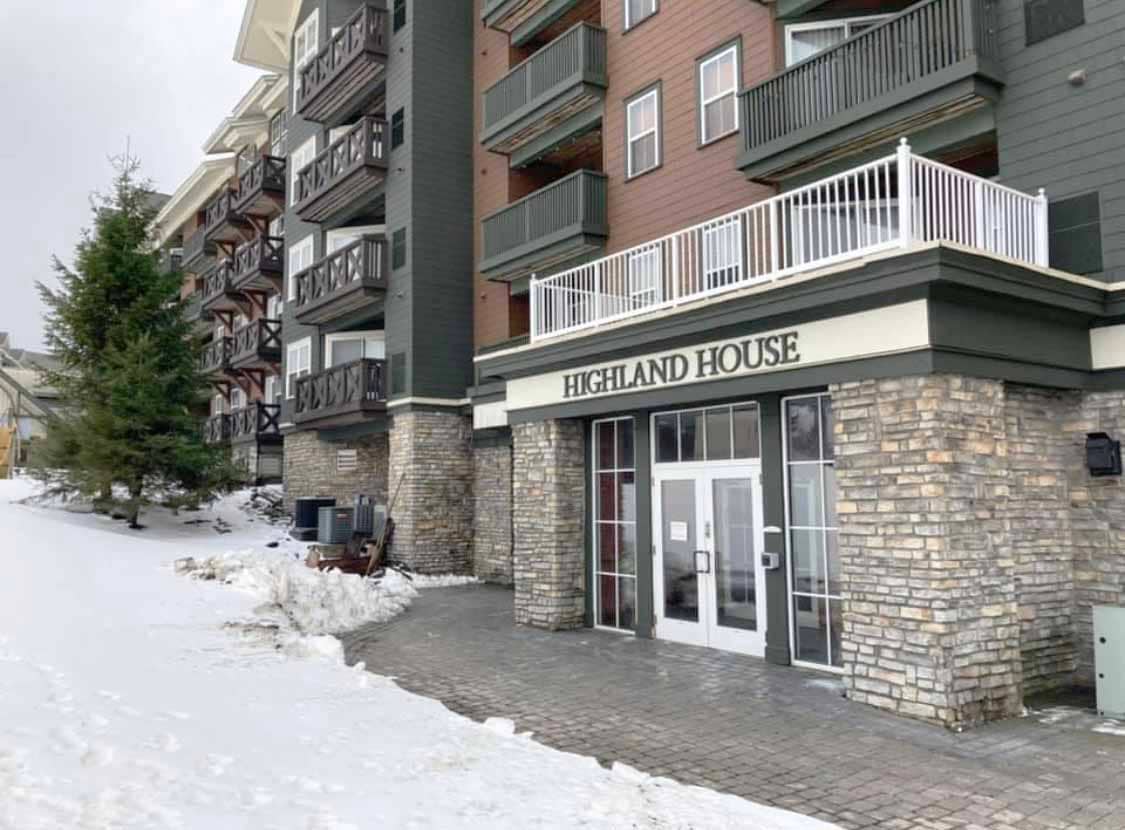 The exterior of the Highland House is displayed, featuring a stone facade and large glass doors. Snow covers the ground, and a spruce tree is visible to the left. The building presents a combination of wood and stone finishes, with balconies accessible from upper floors.