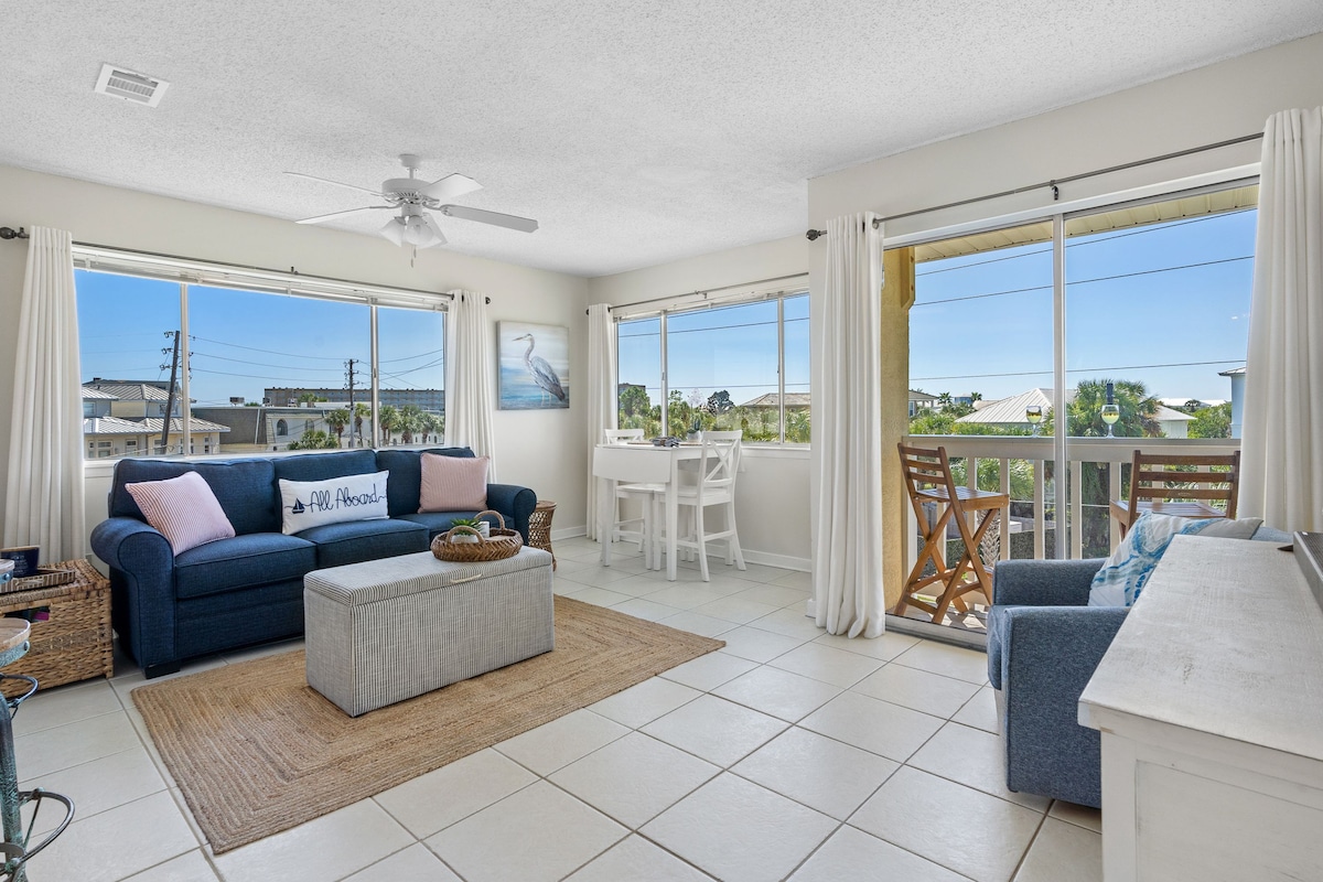 A bright and airy living space features large windows allowing natural light to fill the room, enhancing the views of the outside. A comfortable blue sofa is positioned opposite a light-colored coffee table, with a small dining area placed near the windows.
