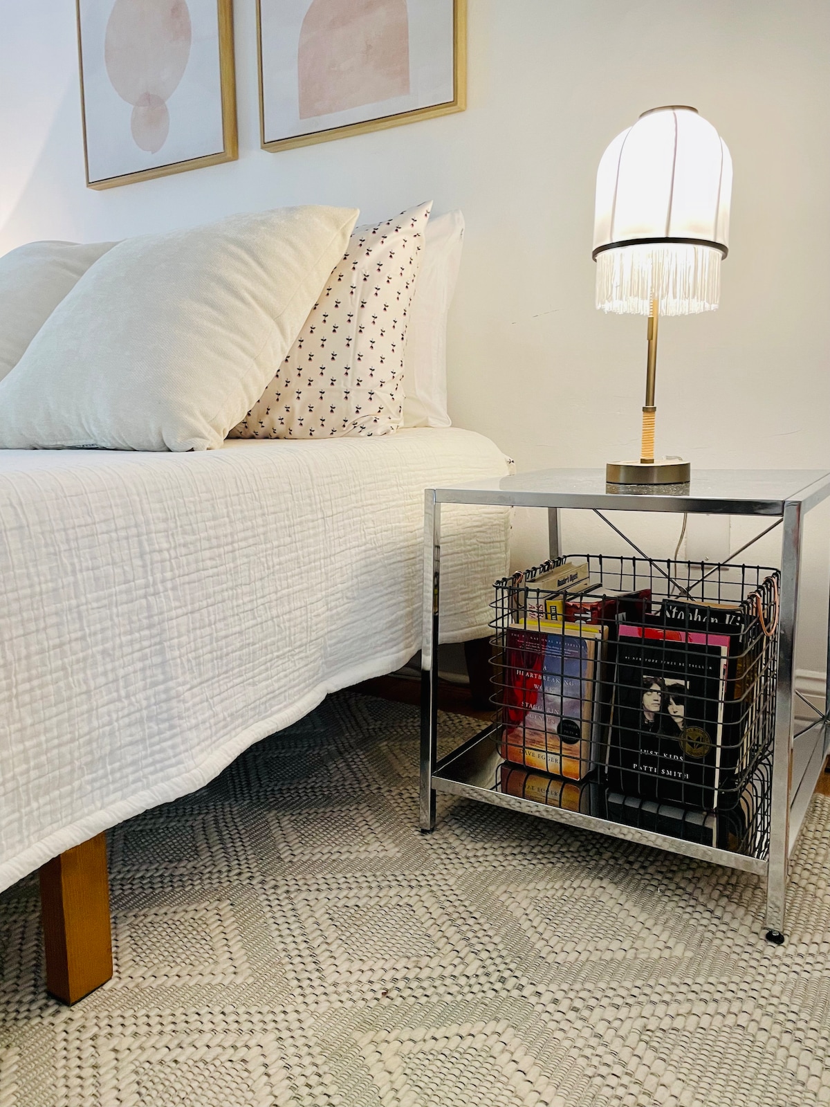 A neatly made bed is showcased with multiple pillows and a light-colored quilt. A modern nightstand features a decorative lamp with a fringe design. Below the table, a wire storage basket holds various items, contributing to the functional design of the sleeping area.