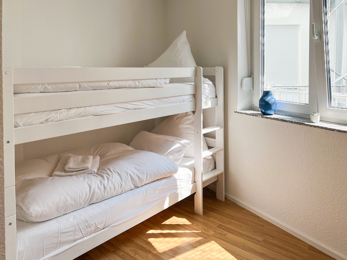 A well-designed sleeping area features a white wooden bunk bed with two comfortable mattresses. Soft white bedding and pillows are neatly arranged. A sunny window allows natural light to illuminate the space, complemented by a decorative blue vase on the window sill.